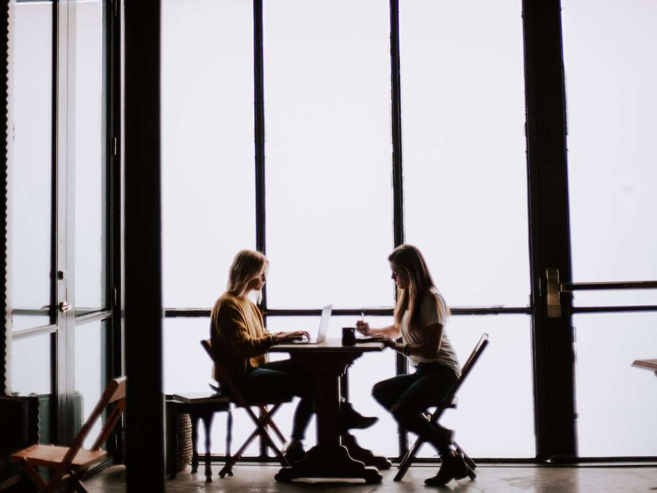 photo of two women facing each other sitting in front of table near glass wall